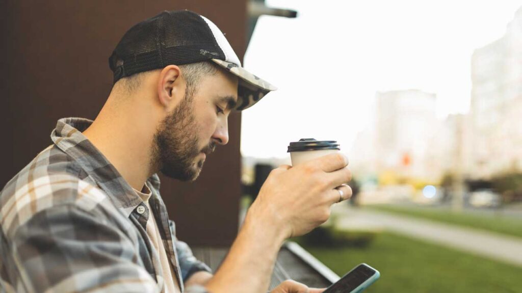 man wearing baseball cap holds coffee and looks at phone